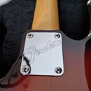 Detail zadní strany kytary s viditelným spojem krku a těla. Na chromovaném krytu je vyryto logo Fender®. Tělo kytary má červenohnatý gradientní lak a…