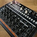 Černý elektronický bicí automat s nápisem DRUMBRUTE IMPACT. Zařízení obsahuje množství bílých a černých otočných ovladačů, několik bílých tlačítek a…