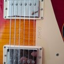 Detail elektrické kytary s červeně-žlutým přechodem barvy korpusu a krémovým pickguardem. Na pickguardu je svisle napsán text 1960. Vidět jsou dva sn…