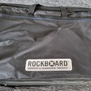 Černý textilní pouzdro na hudební nástroj s dvěma rukojtěmi a zipem. V dolní části je bílý obdélníkový štítek s nápisem ROCKBOARD® a pod ním menší te…