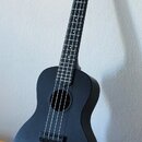 Černý ukulele s matným povrchem. Na hlavě nástroje je bílý nápis „The Waterman USA“. Nástroj vykazuje známky běžného používání a je opřen o bílou zeď…