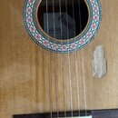 Detail těla akustické kytary s přírodním dřevem. Kolem otvoru je dekorativní rozetový ornament s červenými květy. Uvnitř otvoru je vidět štítek s náp…