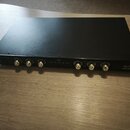 Černý rackový zesilovač s kovovým povrchem. Na předním panelu jsou vlevo tři a vpravo tři zlaté konektory typu XLR. V pravém dolním rohu je bílý nápi…