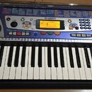 Elektronický klavír v černé barvě s modrým ovládacím panelem a dvěma integrovanými reproduktory po stranách. Na panelu je viditelné logo YAMAHA a ozn…