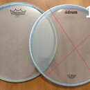 Dvě průhledné bicí kožehy s kovovým obručováním. Levý kožeh nese logo REMO a nápis EMPEROR CLEAR MADE IN USA. Pravý kožeh má v horní části nápis ddru…