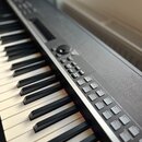 Černý digitální klavír s bílými a černými klávesami. Na ovládacím panelu jsou vidět tlačítka s nápisy A. PIANO, GRY PERC, STRINGS, CHOIR, E. PIANO, P…