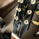 Černá hlava elektrické kytary s nápisy Gibson, Les Paul MODEL a Studio v gold barvě. Nástroj má šest stříbrných ladicích strojů s krémovými knoflíky.…