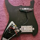 Černý plastový pickguard pro elektrickou kytaru s namontovanou elektronikou a snímači. Na horním snímači je nápis „ALL“ a značka „CE“. Spodní snímač…