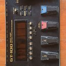 Černý kytarový procesor s nápisem BOSS a GT-100 AMP Effects Processor. Zařízení má displej, několik ovládacích tlačítek a pět stoupakových spínačů s…