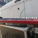 Červený syntetizátor s klávesnicí, umístěný na černém transportním kufru. Na boku je viditelný nápis dwave2 a na zadním panelu jsou různé konektory,…