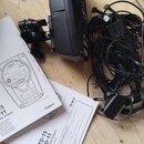 Na dřevěném povrchu je černý elektronický bicí modul s připevněným držákem. Vedle něj leží svazek černých kabelů s napájecím adaptérem a dva papírové…