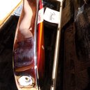 Detailní pohled na bok elektrické kytary s červeně-žlutým přechodem barev (sunburst) v černém kufru. Na kytaru je připevněn bílý papírek s nápisem Fe…