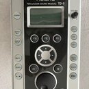 Stříbročerný perkusní modul s displejem a ovládacími prvky. Na horní části je bílý nápis Roland a pod ním text PERCUSSION SOUND MODULE TD-9. V dolní…