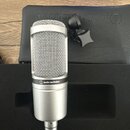 Stříbrný kondenzátorový mikrofon s kovovou síťkou v horní části. Na těle je umístěno logo a nápis audio-technica. Mikrofon leží v černém polstrování…