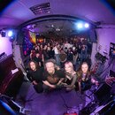 Širokoúhlá fotografie zachycující skupinu lidí v interiéru s fialovým osvětlením. V popředí klečí čtyři osoby, z nichž jedna drží elektrickou basovou…