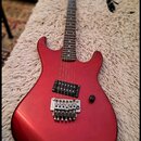Elektrická kytara s červeným metalickým povrchem korpusu a hnědým hmatníkem. Na kobylce je viditelný nápis Floyd Rose. Nástroj má jeden humbucker v p…