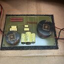 Elektronický obvod na zelené desce umístěný na černém plátně. Na desce jsou dva velké černé transformátory, několik kondenzátorů a rezistory. V horní…