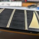 Detailní pohled na hриф nástroje s černým hmatníkem a kovovými pražci. Na hmatníku jsou vidět světlé trojúhelníkové vložky (inlaye). Povrch hmatníku…