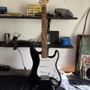 Černá elektrická kytara s bílým pickguardem a hnědým hnedobarevným hmatníkem. Na hlavě nástroje je viditelné logo Squier. Kytara stojí na černém stoj…