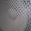 Detailní záběr na kovovou perforovanou mřížku stříbrné barvy. Mřížka tvoří pravidelnou strukturu z oválných otvorů. Na povrchu nejsou vidět žádné zna…
