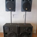 Dva černé reproduktorové sety sestávající z horních satelitů a spodních basových beden na stojaných tyčích. Horní reproduktory mají v horní části roh…