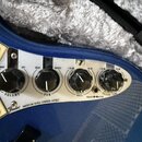 Detail ovládacího panelu modré elektrické kytary s bílým pickguardem. Na panelu jsou čtyři černé otočné knoflíky s nápisy VOLUME, PAN, TREBLE BASS a…
