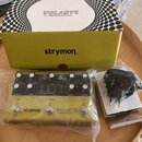 Na fotografii je efekt Volante značky Strymon v originální krabici s přiloženým napájecím adaptérem. Pedál má olivově zelený povrch a nápisy „strymon…
