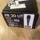 Fotografie znázorňuje černou kartonovou krabici od mikrofonu. Na čelní straně je bílý nápis PR 20 UT, pod ním slovo Black a seznam příslušenství: Inc…