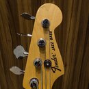 Fotografie zachycuje hlavu basové kytary z přírodního světlého dřeva s chromovanými ladičkami. Na povrchu je černý nápis Fender® JAZZ BASS. Povrch dř…