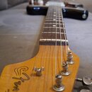 Fotografie zachycuje hmatník a hlavu elektrické kytary z vybarveného dřeva. Na hlavě je černé logo "Fender". Nástroj má šest kovových ladiček a tmavý…