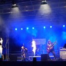Fotografie zachycuje hudební kapelu během koncertu na scéně s modrým osvětlením a kouřem. V popředí jsou vidět dva hudebníci, z nichž jeden hraje na…