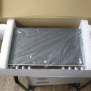 Černý elektronický přístroj v kartonové krabici s bílými polystyrenovými vložkami. Zařízení je zabaleno v průhledné plastové fólii, která zakrývá hor…