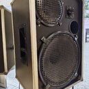 Černý reproduktor v hranatém kufrovém provedení s kovovými hranami. Přední strana obsahuje dva reproduktory chráněné mřížkami a jeden menší tubový re…