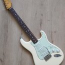 Bílá elektrická kytara s jasně viditelným logem Fender na hlavě. Nástroj má tmavý hmatník, světle zelený pickguard a tři snímače. Povrch kytary vypad…