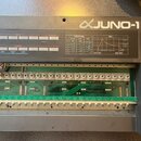 Šedý syntetizátor s nápisem JUNO-1. Horní panel obsahuje tabulky WAVEFORM a ENV oraz ovládací prvky BANK a NUMBER. Přístroj je otevřený, vnitřní pros…