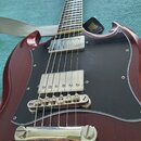 Červená elektrická kytara s černým pickguardem a dvěma snímači s kovovými kryty. Na černé popruhu je viditelné logo ERNIE BALL. Povrch kytary vykazuj…