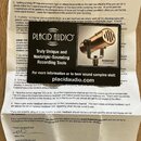 Fotografie zobrazuje printed manuál s textem v angličtině. Uprostřed je umístěn černý reklamní leták značky PLACID AUDIO s nápisem Truly Unique and N…