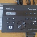 Černý elektronický bicí modul s nápisy Roland a V-Drums. Na displeji je zobrazen text DRUM KIT 01 LiveAcoustic a v pravém dolním rohu je modelové ozn…