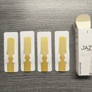 Čtyři trostové plátky pro saxofon v bílých papírových obalů. Vedle nich je umístěna část bílé krabičky s nápisy JAZZ, 5 UNFILED a TENOR SAXOPHONE. V…