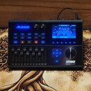 Černý elektronický bicí modul s displejem a ovládacími prvky. Na levé straně je modré logo ALESIS, vpravo dole bílý nápis STRIKE a pod ním menší text…