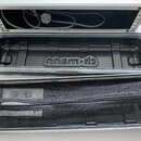 Otevřený hliníkový kufřík s černým plastovým vnitřním luskem. Na dně je vyraženo logo „km-h“. V horní části kufříku je srolovaný napájecí kabel a v p…