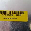 Detail kovového povrchu s nalepenou žlutou štítkem. Štítek obsahuje čárový kód, číslo 7710515 - 000 a nápis PG ASSY AM PRO STRAT PBP. Povrch kovu vyk…