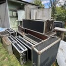 Skupina přepravních kufrů (flightcasů) různých velikostí v černé a stříbrné barvě, umístěná venku na trávě. Kufry mají kovové hrany a rohové zpevnění…