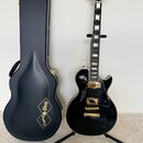 Černá elektrická kytara s gold hardwarem na stojanu, černý tvrdý kufýr a zlatý instrumentální kabel s tunerem. Na hlavě kytary je nápis Epiphone a na…
