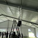 Interiér bílého stanu s kovovou konstrukcí. Uprostřed je umístěn vertikální hliníkový truss, na kterém visí černý reproduktor. V popředí stojí hliník…