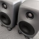 Dva šedé studiové monitory s matným povrchem a zaoblenými hranami. Na spodní části obou reproduktorů je bílý nápis GENELEC. Každý monitor má v horní…