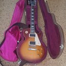 Elektrická kytara s tónem sunburst v hnědých a žlutých odstínech, bílým pickguardem a dvěma humbuckery. Nástroj je umístěn v otevřeném hnědém tvrdém…