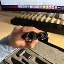Detailní pohled na konce dvou černých trubic状ých komponentů, pravděpodobně částí hudebního nástroje nebo příslušenství. Levý konec je otevřený a hlub…