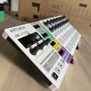 Bílý sekvencer s černými ovládacími prvky a barevnými tlačítky. Na horní části je nápis ARTURIA YOUR EXPERIENCE YOUR SOUND. V pozadí je vidět kartono…