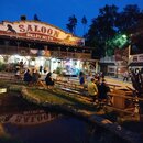 Fotografie zobrazuje dřevěnou budovu v stylu divadla nebo saloonu s nápisem SALOON ŠIKLŮV MLÝN a číslem 1899. Před budovou se nachází venkovní terasa…